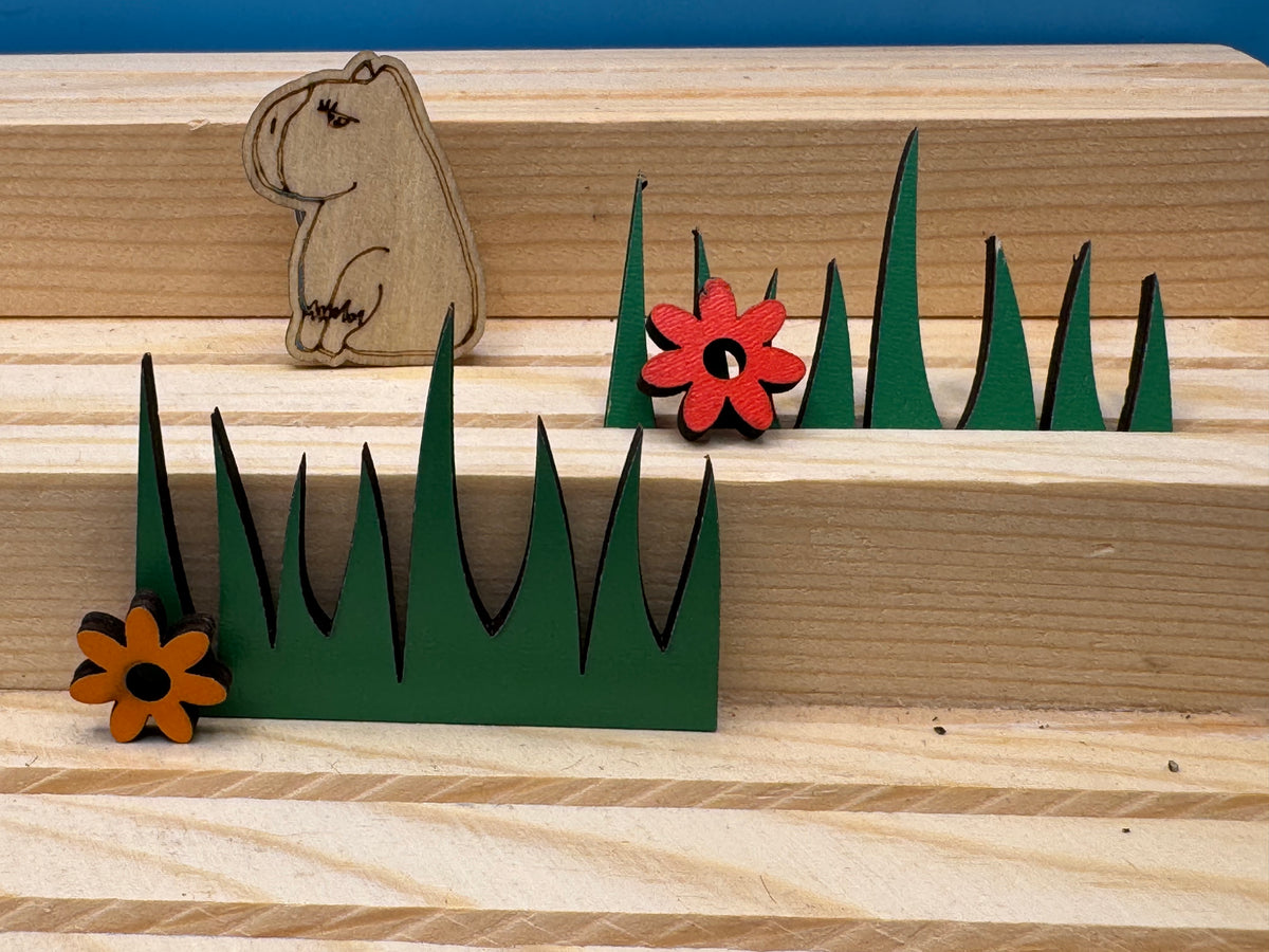 A hand drawn wooden capybara pin badge, arranged on a light wood surface with flowers.

