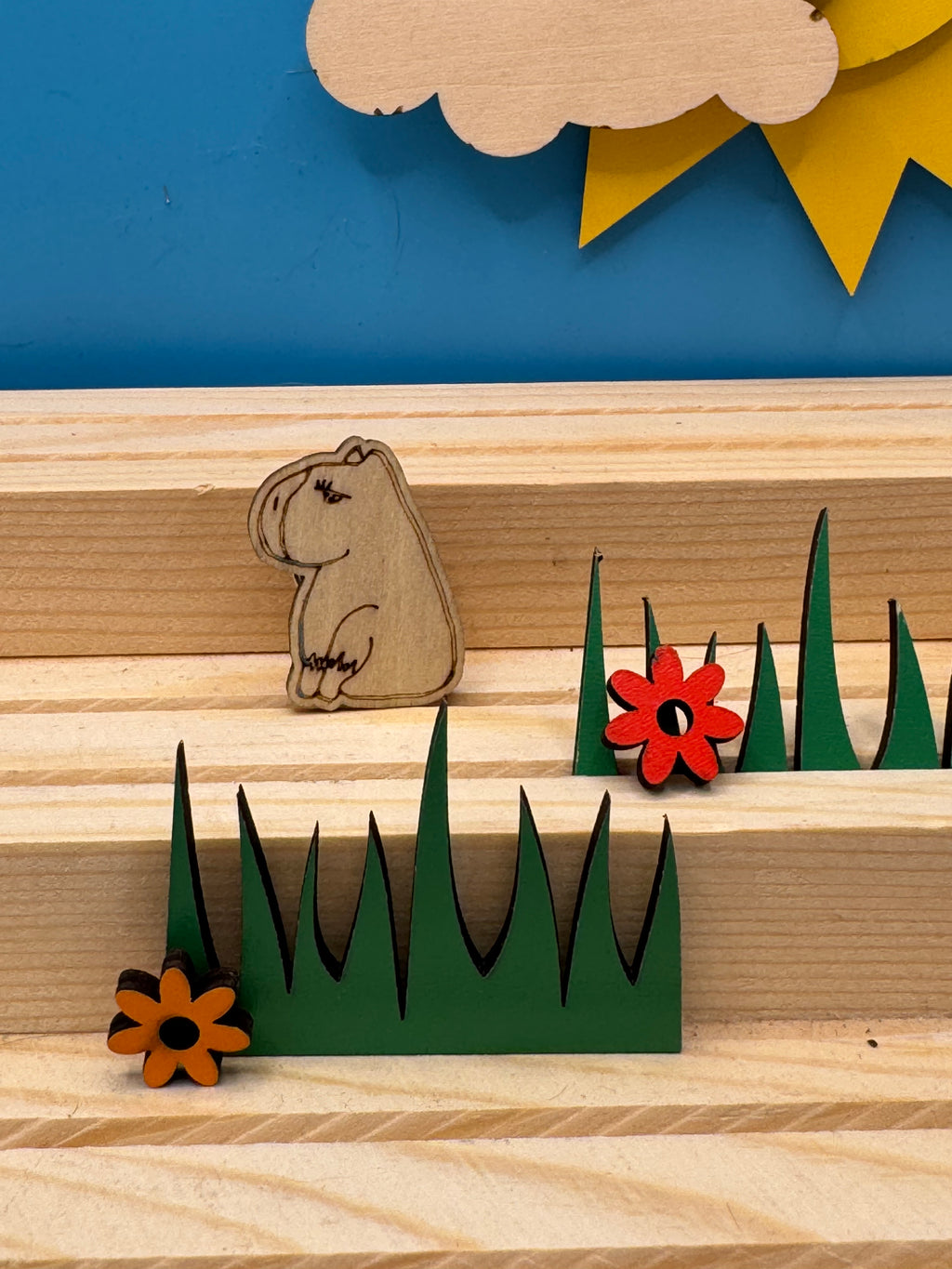 A hand drawn wooden capybara pin badge sitting on a green tiered landscape with two small wooden flowers, displayed on a light wood striped surface against a blue background.
