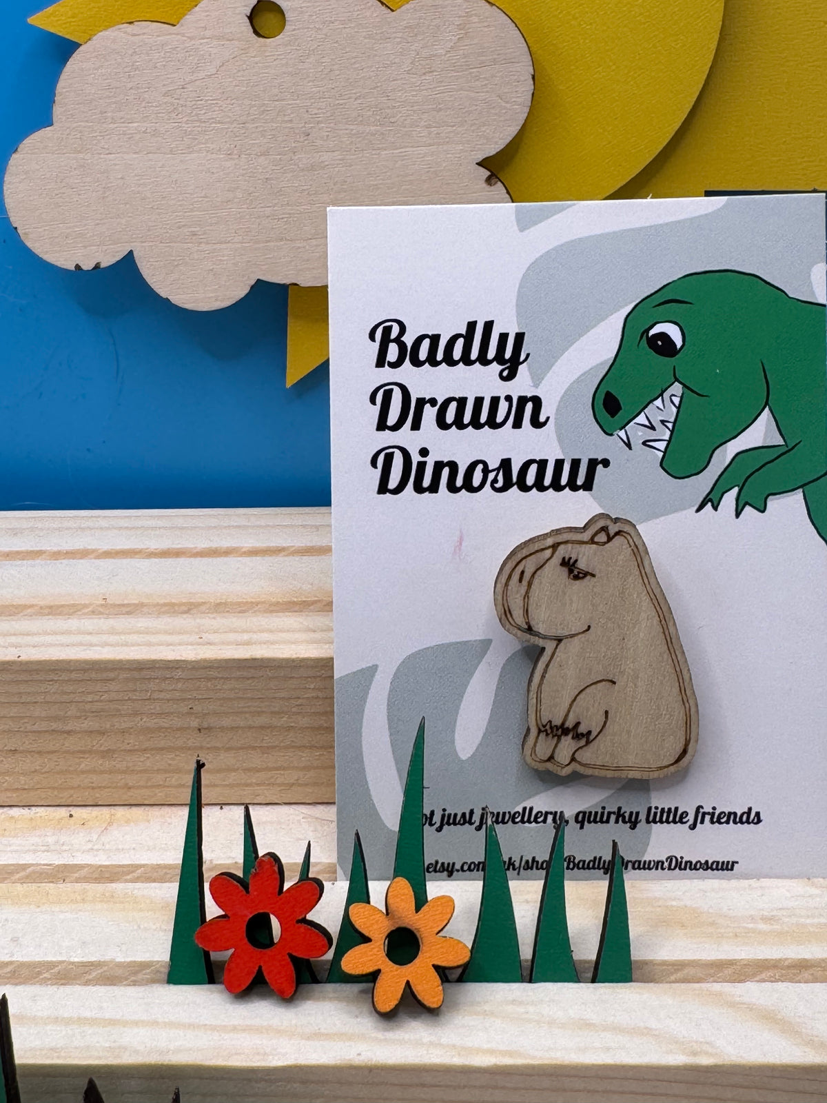 A hand drawn wooden capybara pin badge sitting on a green tiered landscape with two small wooden flowers, displayed on a light wood striped surface against a blue background.
