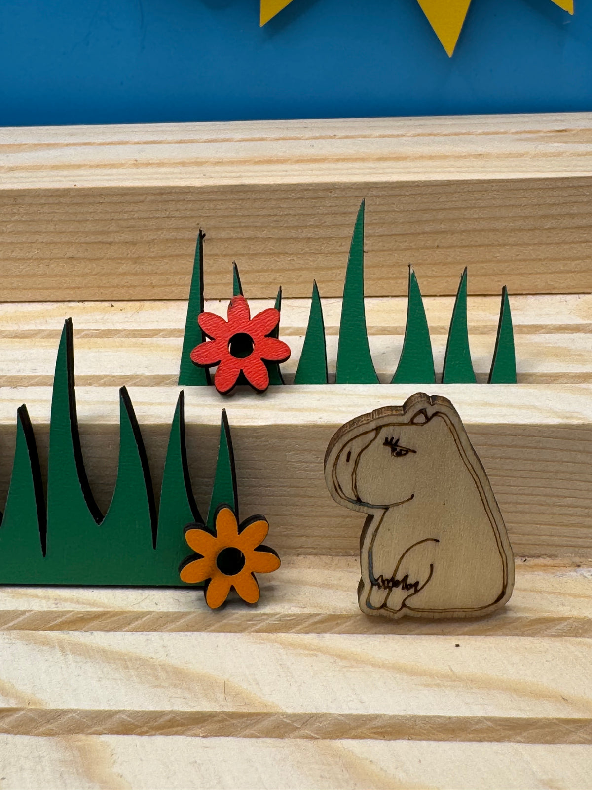 A hand drawn wooden capybara pin badge sitting on a green tiered landscape with two small wooden flowers, displayed on a light wood striped surface against a blue background.
